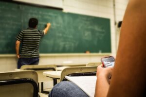 En un aula iluminada por luz natural, un profesor está de espaldas a la cámara, escribiendo en la pizarra verde. Un estudiante en primer plano está sentado, aparentemente distraído por su teléfono móvil en lugar de prestar atención a la clase o sus notas, simbolizando el desafío de involucrar a los alumnos en la era de la distracción digital.