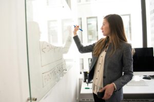 Una mujer profesional está escribiendo en una pizarra blanca transparente en una oficina luminosa con ventanas grandes. Ella parece estar esbozando un diseño o una estructura, con palabras como "FOOTER" visibles, lo que sugiere que podría estar planificando un layout de sitio web o una interfaz de usuario. Su atuendo y la disposición de la sala sugieren un entorno de trabajo colaborativo y educativo donde las habilidades blandas, como la comunicación y la planificación, son importantes.