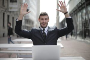 Un hombre de negocios sonriente levanta las manos en gesto de éxito frente a su portátil en la calle.