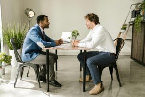 Dos hombres de negocios se sientan en una mesa, participando en una conversación animada, probablemente evaluando habilidades blandas en un contexto profesional.